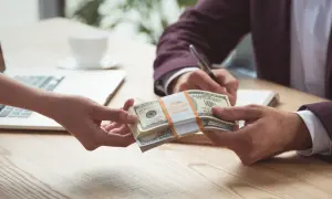 Dwie ręce przekazują sobie stos banknotów dolarowych na tle laptopa i filiżanki kawy.