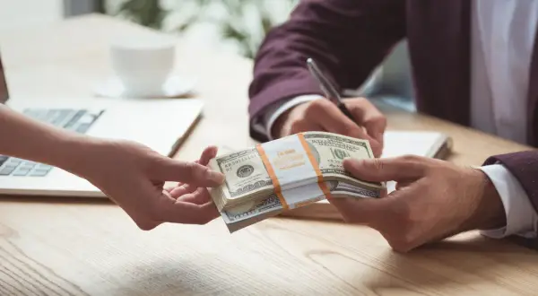 Dwie ręce przekazują sobie stos banknotów dolarowych na tle laptopa i filiżanki kawy.