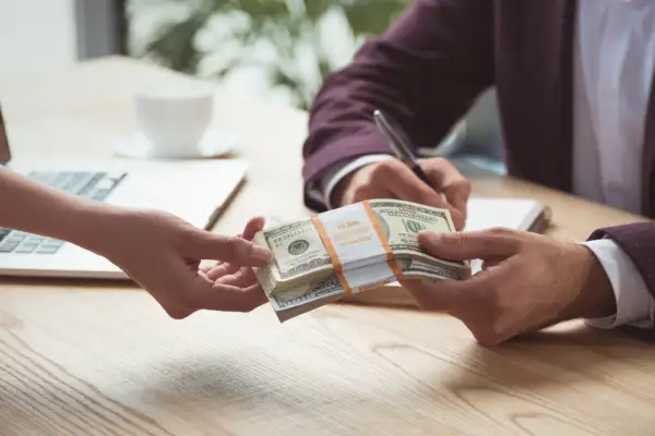 Dwie ręce przekazują sobie stos banknotów dolarowych na tle laptopa i filiżanki kawy.
